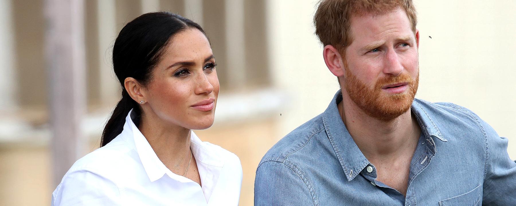 Meghan sitzt mit weißer Bluse und Zopf-Frisur neben Harry im hellblauen Jeanshemd auf einer Bank.