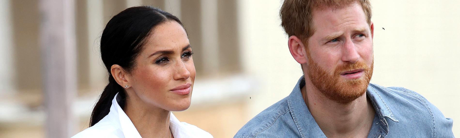 Meghan sitzt mit weißer Bluse und Zopf-Frisur neben Harry im hellblauen Jeanshemd auf einer Bank.