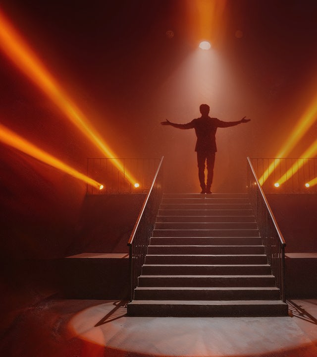 Eine einzelne Person steht oben auf einer breiten Treppe auf einer dunklen Bühne, die Arme weit ausgebreitet. Von hinten und von den Seiten strahlen mehrere orangefarbene Scheinwerfer durch Nebel und erzeugen eine dramatische Lichtstimmung. Die Person ist nur als Silhouette zu erkennen, während die Treppe und das Bühnenlicht im Vordergrund deutlich sichtbar sind.