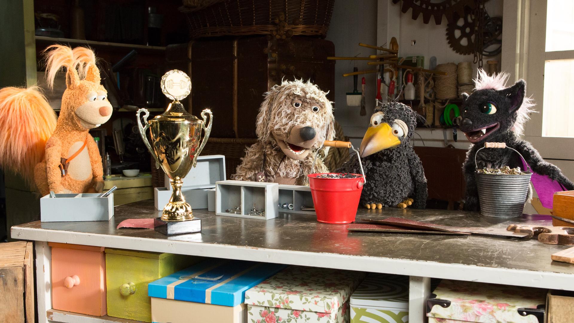 udis Rasselbande ist in der Garage um Rudi herum versammelt und schaut ihn angespannt an. Rudi greift mit dem Schnabel den Henkel eines vollen Eimers. Vor ihnen steht ein großer Pokal auf dem Tisch. 