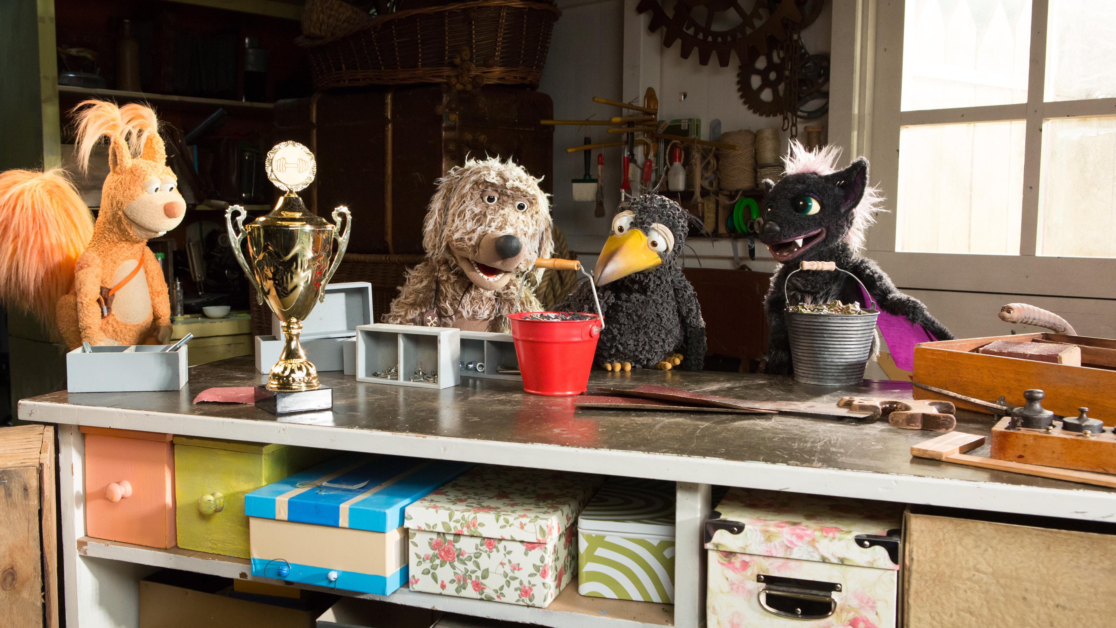 udis Rasselbande ist in der Garage um Rudi herum versammelt und schaut ihn angespannt an. Rudi greift mit dem Schnabel den Henkel eines vollen Eimers. Vor ihnen steht ein großer Pokal auf dem Tisch. 