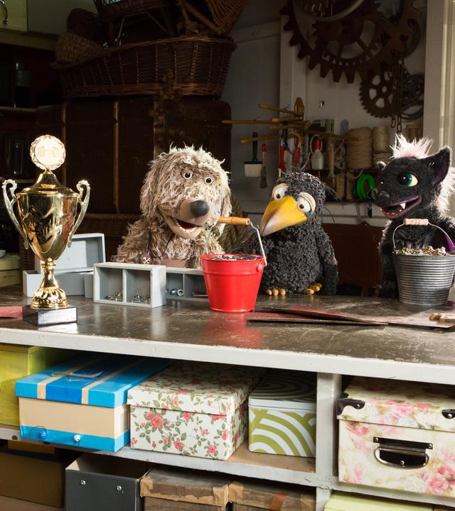 udis Rasselbande ist in der Garage um Rudi herum versammelt und schaut ihn angespannt an. Rudi greift mit dem Schnabel den Henkel eines vollen Eimers. Vor ihnen steht ein großer Pokal auf dem Tisch. 