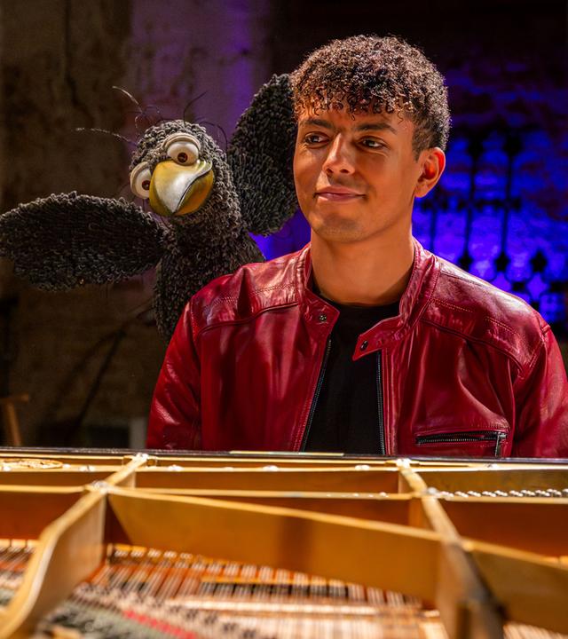 Blick durch den Flügel: Louis Philippson und Rudi sitzen hinter dem Flügel und schauen freudig in die Kamera. Rudi hebt dabei seine Flügel.