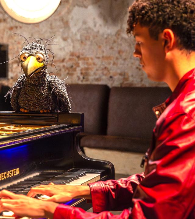 Blick durch den Flügel: Louis Philippson und Rudi sitzen hinter dem Flügel und schauen freudig in die Kamera. Rudi hebt dabei seine Flügel.
