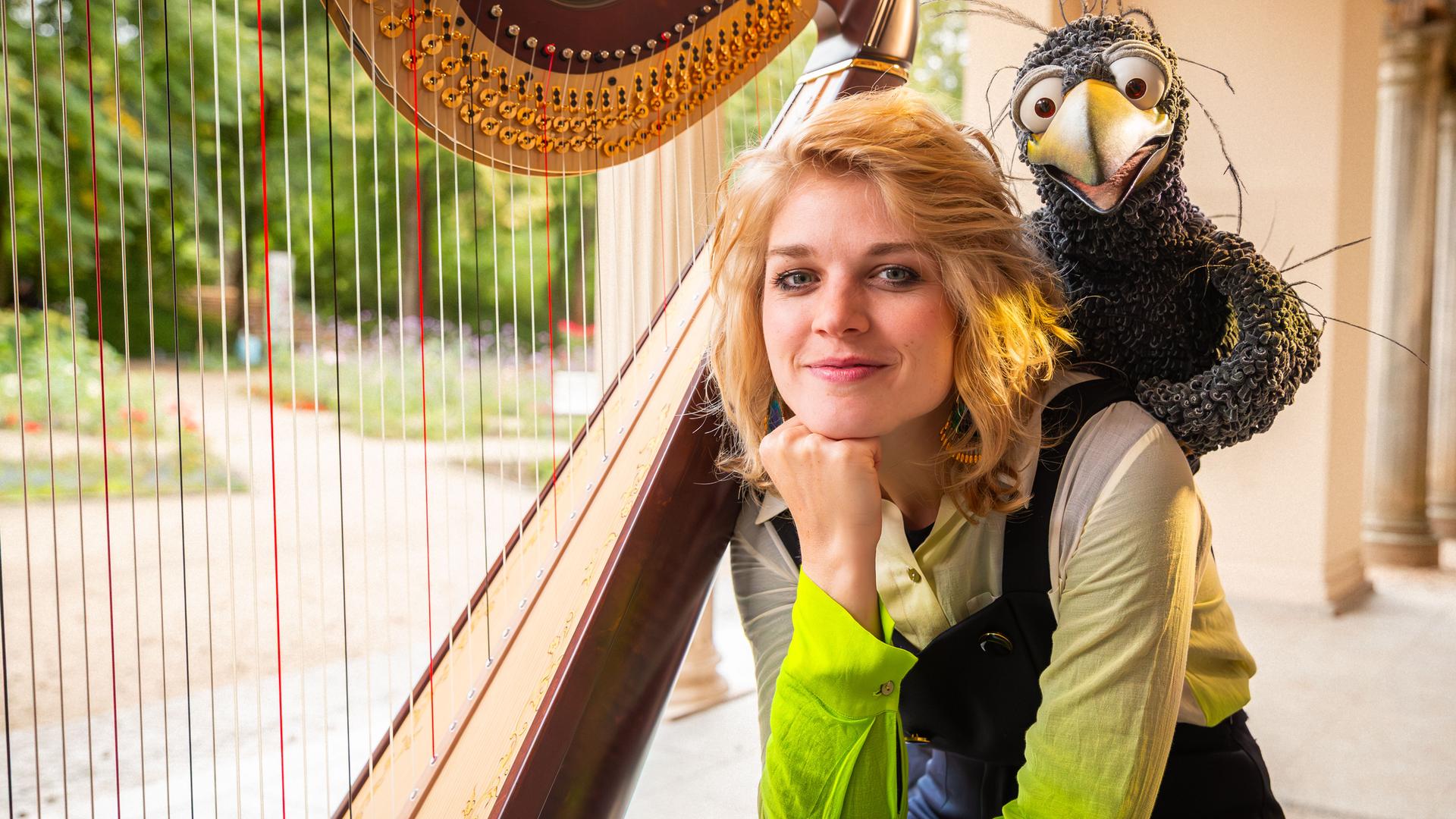 Rabe Rudi hockt auf der Schulter von Magdalena Hoffmann. Beide schauen freudig in die Kamera, neben ihnen steht eine Harfe.