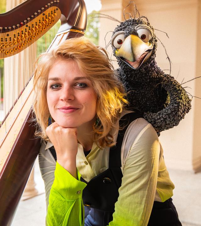 Rabe Rudi hockt auf der Schulter von Magdalena Hoffmann. Beide schauen freudig in die Kamera, neben ihnen steht eine Harfe.