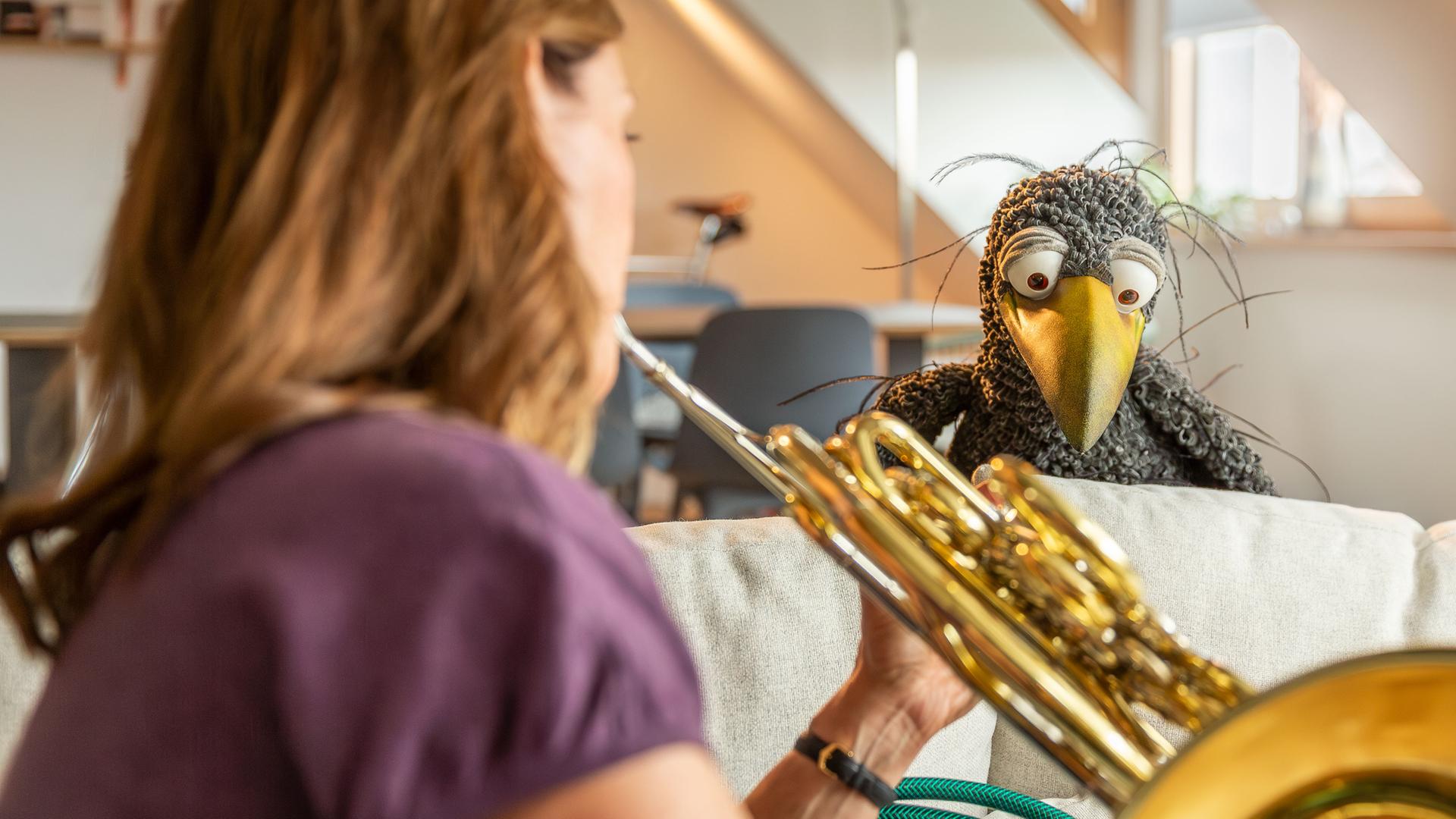 Wir sehen Sarah Willis von hinten im Anschnitt, die ihr Horn bläst. Vor ihr auf der Sofalehne hockt Rudi und schaut ihr neugierig zu.