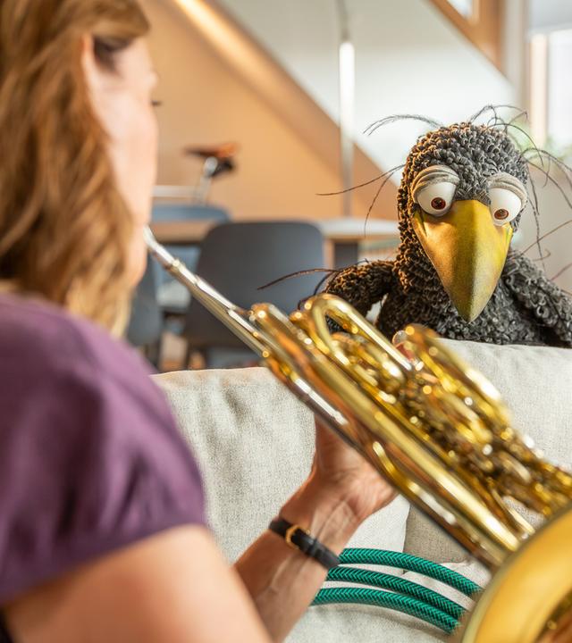 Wir sehen Sarah Willis von hinten im Anschnitt, die ihr Horn bläst. Vor ihr auf der Sofalehne hockt Rudi und schaut ihr neugierig zu.