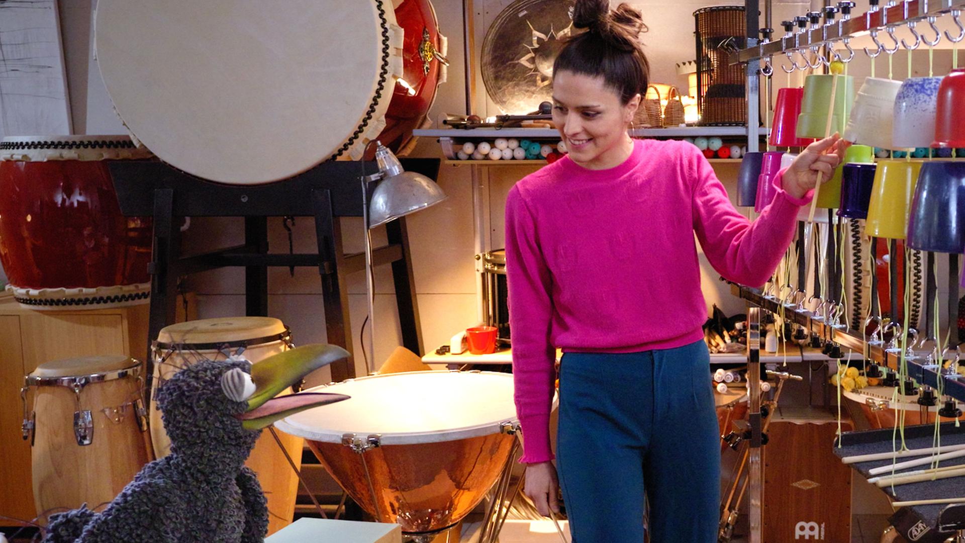 Rudi und Vivi Vassileva sind in einem Raum voller Percussion-Instrumente und schauen sich an. Vivi hat zwei Schlagstöcke in den Händen und steht vor ihrem „Blumentopfophon“, einer Reihe aufgehängter Blumentöpfe.