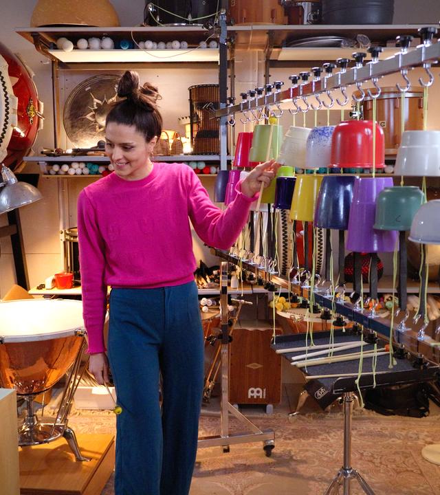 Rudi und Vivi Vassileva sind in einem Raum voller Percussion-Instrumente und schauen sich an. Vivi hat zwei Schlagstöcke in den Händen und steht vor ihrem „Blumentopfophon“, einer Reihe aufgehängter Blumentöpfe.