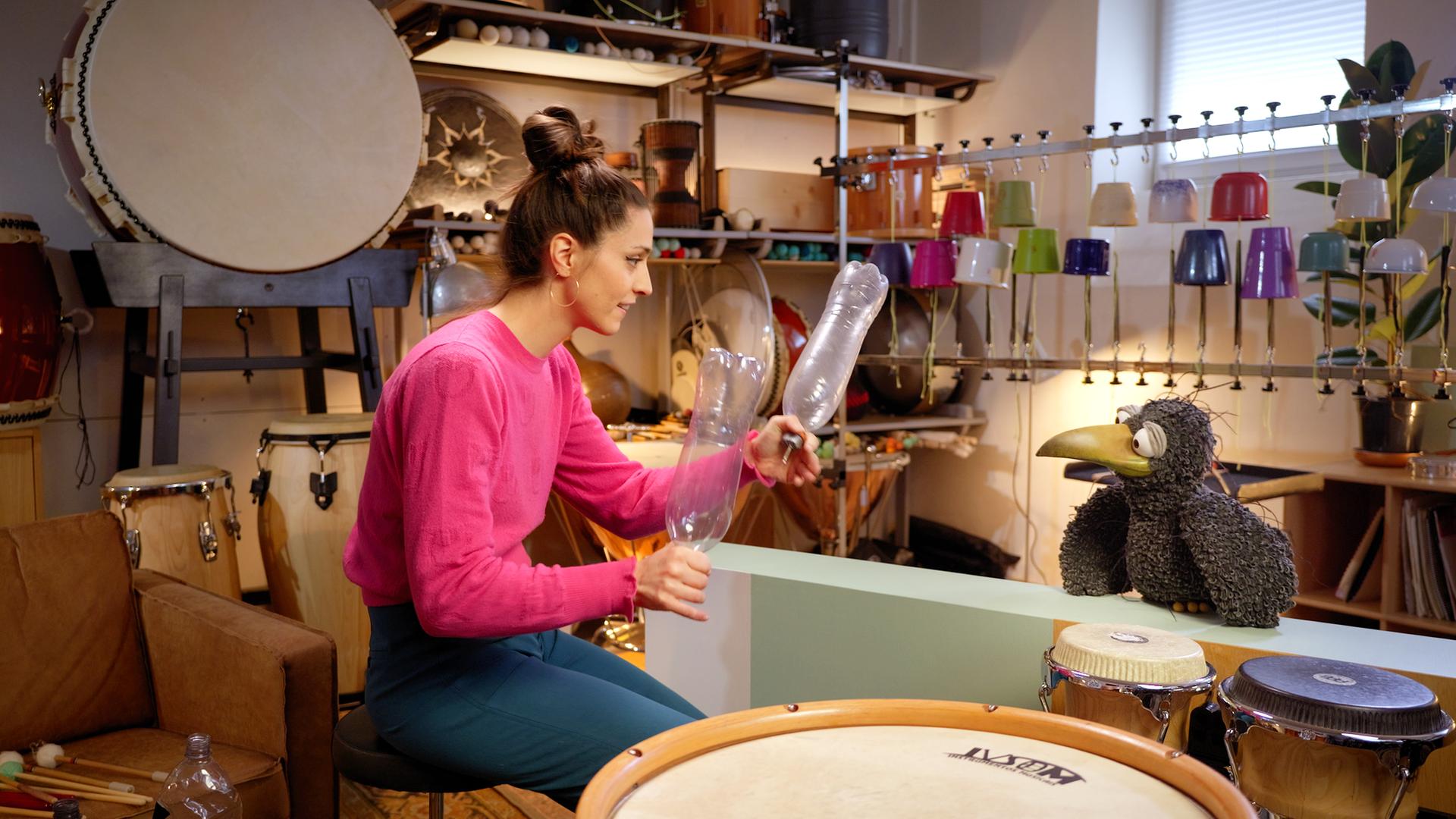 Vivi Vassileva und Rudi sind in einem Raum voller Percussion-Instrumente. Rudi schaut zu Vivi, die vor ihm sitzt und zwei leere Plastikflaschen in den Händen hält. 