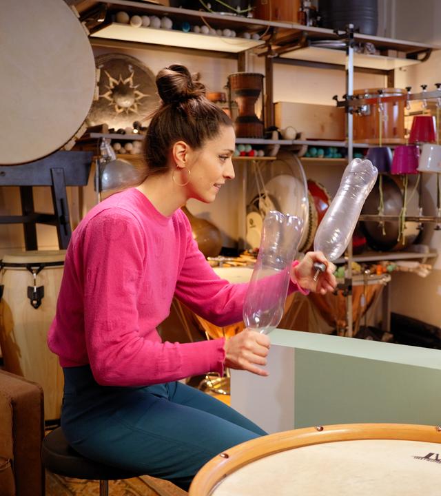 Vivi Vassileva und Rudi sind in einem Raum voller Percussion-Instrumente. Rudi schaut zu Vivi, die vor ihm sitzt und zwei leere Plastikflaschen in den Händen hält. 