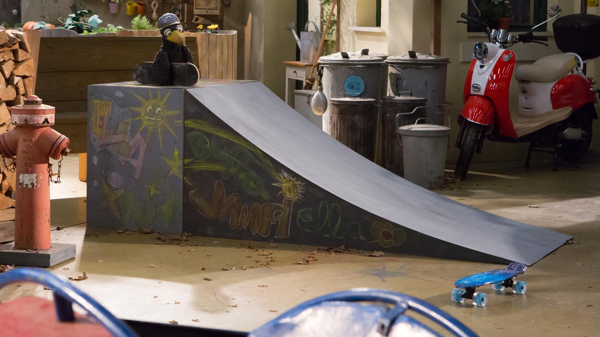 Rudi hockt mit seinem Skateboard und einem Helm auf dem Kopf oben auf einer Rampe und schaut sehnsüchtig auf ein schickes, blaues Board, das unten am Boden liegt.