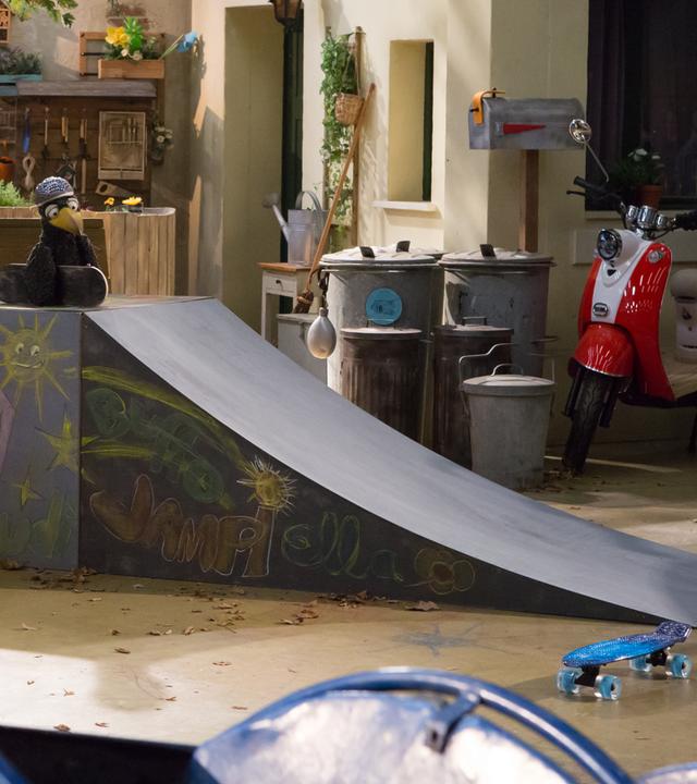 Rudi hockt mit seinem Skateboard und einem Helm auf dem Kopf oben auf einer Rampe und schaut sehnsüchtig auf ein schickes, blaues Board, das unten am Boden liegt.