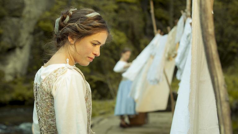 "Rübezahls Schatz": Rosa (Henriette Confurius) mit hochgestecktem Haar, weißer Bluse und geschnürtem Arbeitskleid. Sie steht nachdenklich vor einer Wäscheleine, auf der frisch gewaschene Laken zum Trocknen aufgehängt sind.