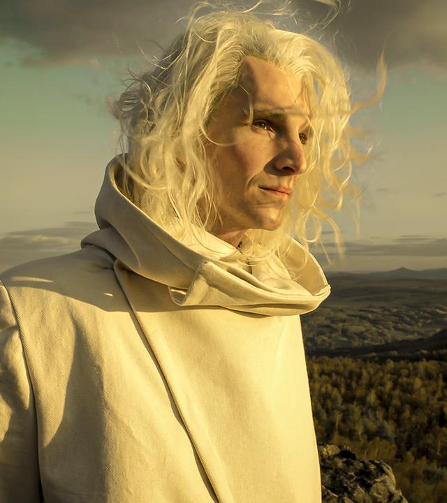 "Rübezahls Schatz": Im Hintergrund eine sich bis zum Horizont erstreckende Waldlandschaft. Im Vordergrund Rübezahl (Sabin Tambrea). Er hat langes, blondes, im Wind wehendes Haar und trägt einen weißen Mantel mit breiten Kragen. Ernst schaut er ins Sonnenlicht.