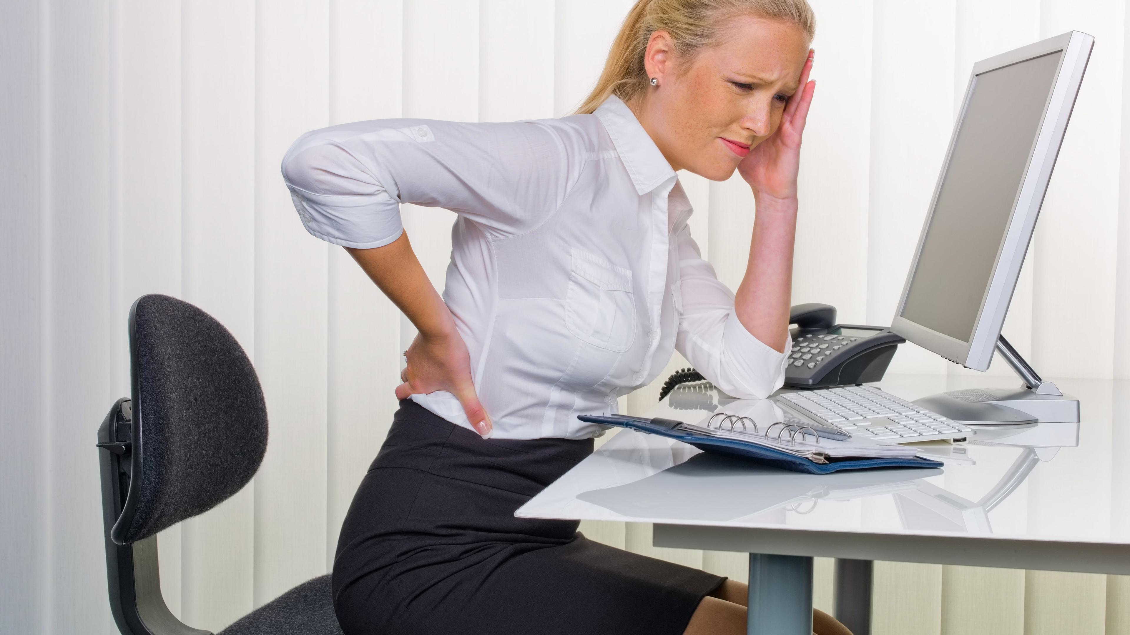 Eine Frau sitzt an einem Schreibtisch und hält sich den Rücken.