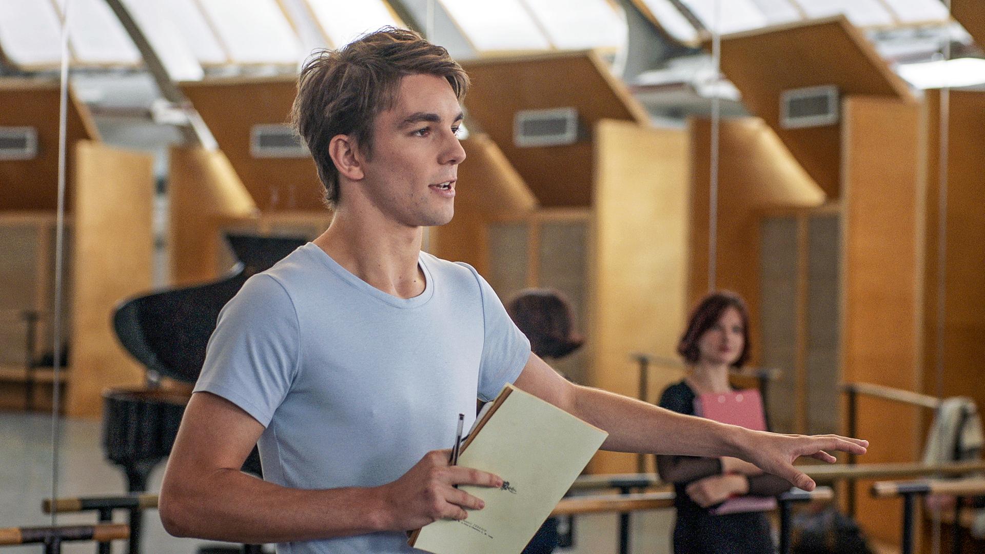 Max steht im Probenraum und hält mir seiner rechten Hand ein Notizbuch fest. Hinter ihm ist eine verspiegelte Wand.