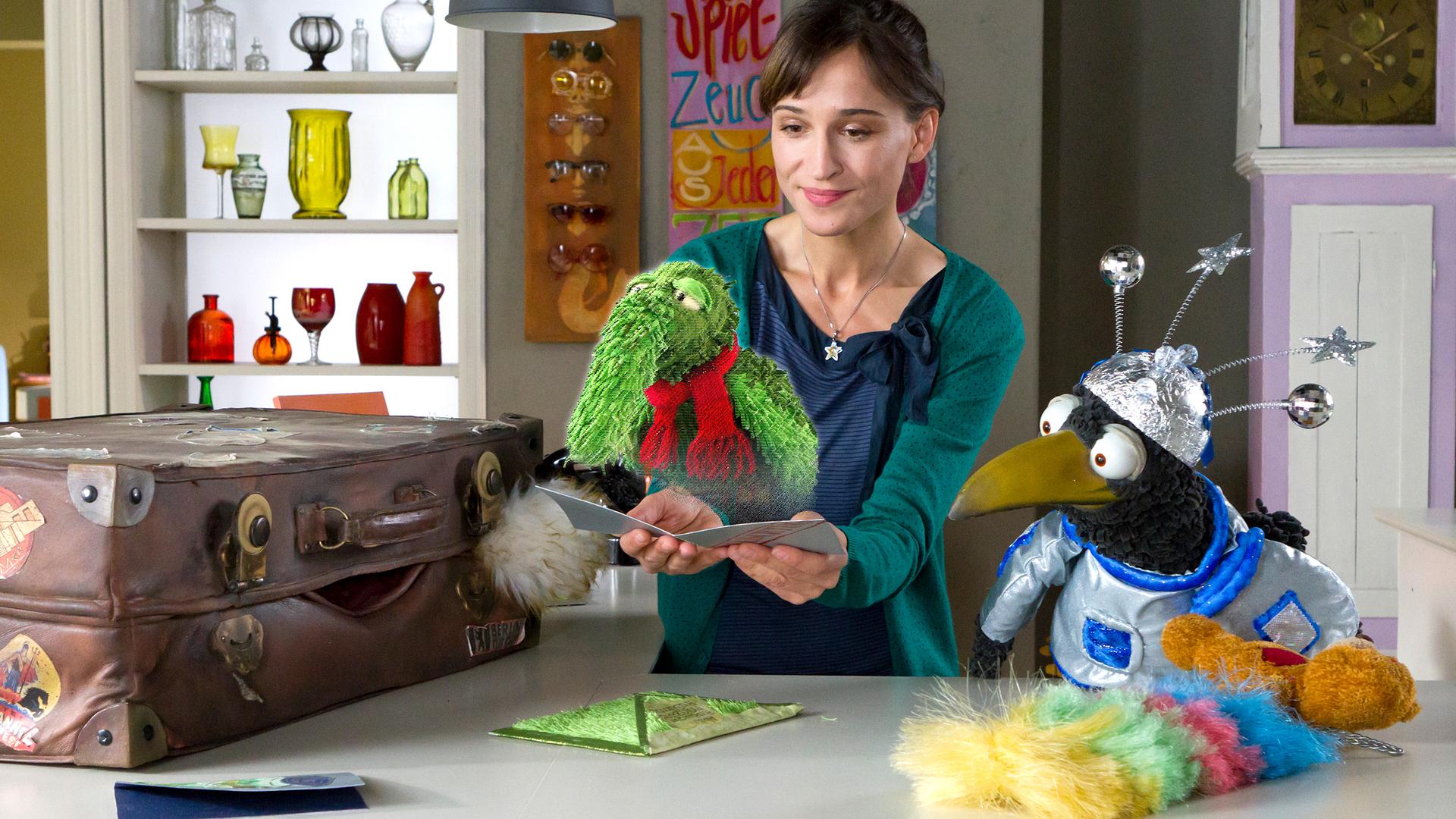 Siebenstein hält eine Karte, auf der die Projektion eines grünen Wesens mit Rüssel, ihrem Freund Rüdügür, zu sehen ist. Der Koffer und Rabe Rudi hocken neben ihr und schauen zu Rüdügür.