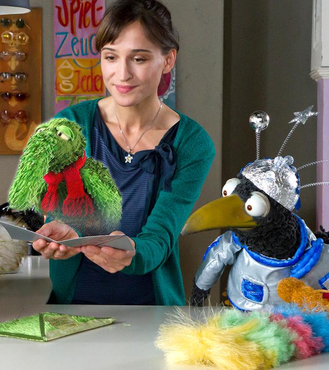 Siebenstein hält eine Karte, auf der die Projektion eines grünen Wesens mit Rüssel, ihrem Freund Rüdügür, zu sehen ist. Der Koffer und Rabe Rudi hocken neben ihr und schauen zu Rüdügür.