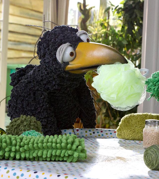 Rabe Rudi hockt neben Rüdügür, einem grünen Wesen mit Rüssel, und reicht ihm mit dem Schnabel einen hellgrünen Schwamm. Auf dem Küchentisch liegen noch weitere grüne Gegenstände verstreut.