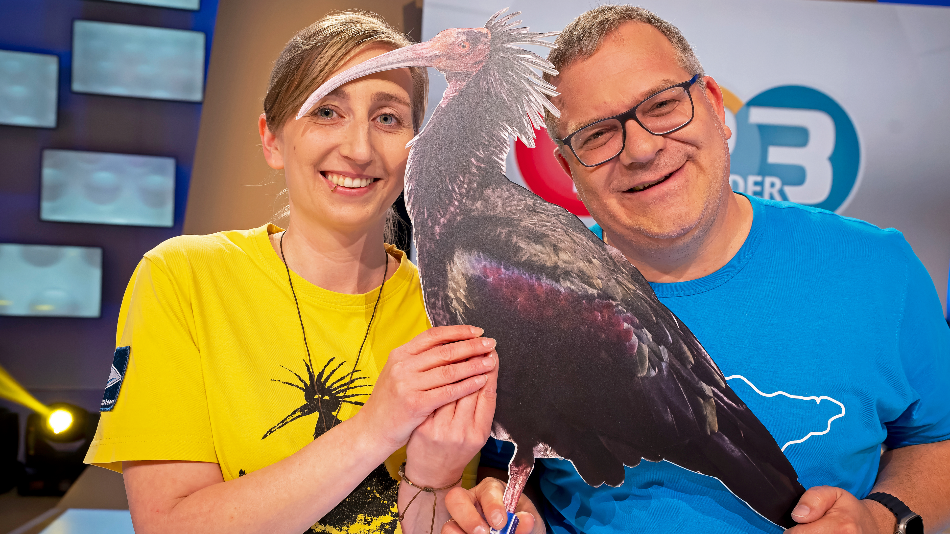Elton und Anne-Gabriele Schmalstieg stehen nebeneinander. Anne-Gabriele trägt ein gelbes T-Shirt, Elton ein blaues. Sie halten ein lebensgroßes Bild von einem Waldrapp in den Händen.