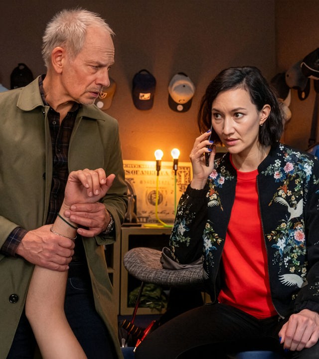 "SOKO Potsdam - Running Dinner": In einem kleinen, abgedunkelten Raum stehen die Ermittler Bernhard Henschel (Michael Lott) und Pauline Hobrecht (Agnes Decker) dicht beieinander. Bernhard hält mit konzentriertem Blick den Unterarm einer Person und prüft den Puls. Pauline sitzt angespannt daneben, telefoniert und hört aufmerksam zu, bereit Anweisungen weiterzugeben.