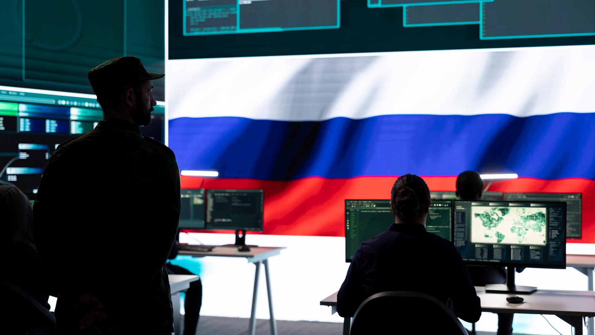 In einem Raum mit einem riesigen Bildschirm, auf dem Daten und eine russische Flagge zu sehen sind, sitzen Personen vor Bildschirmen. Hinter ihnen steht ein Mann in Militäruniform und schaut ihnen zu.