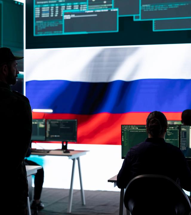 In einem Raum mit einem riesigen Bildschirm, auf dem Daten und eine russische Flagge zu sehen sind, sitzen Personen vor Bildschirmen. Hinter ihnen steht ein Mann in Militäruniform und schaut ihnen zu.