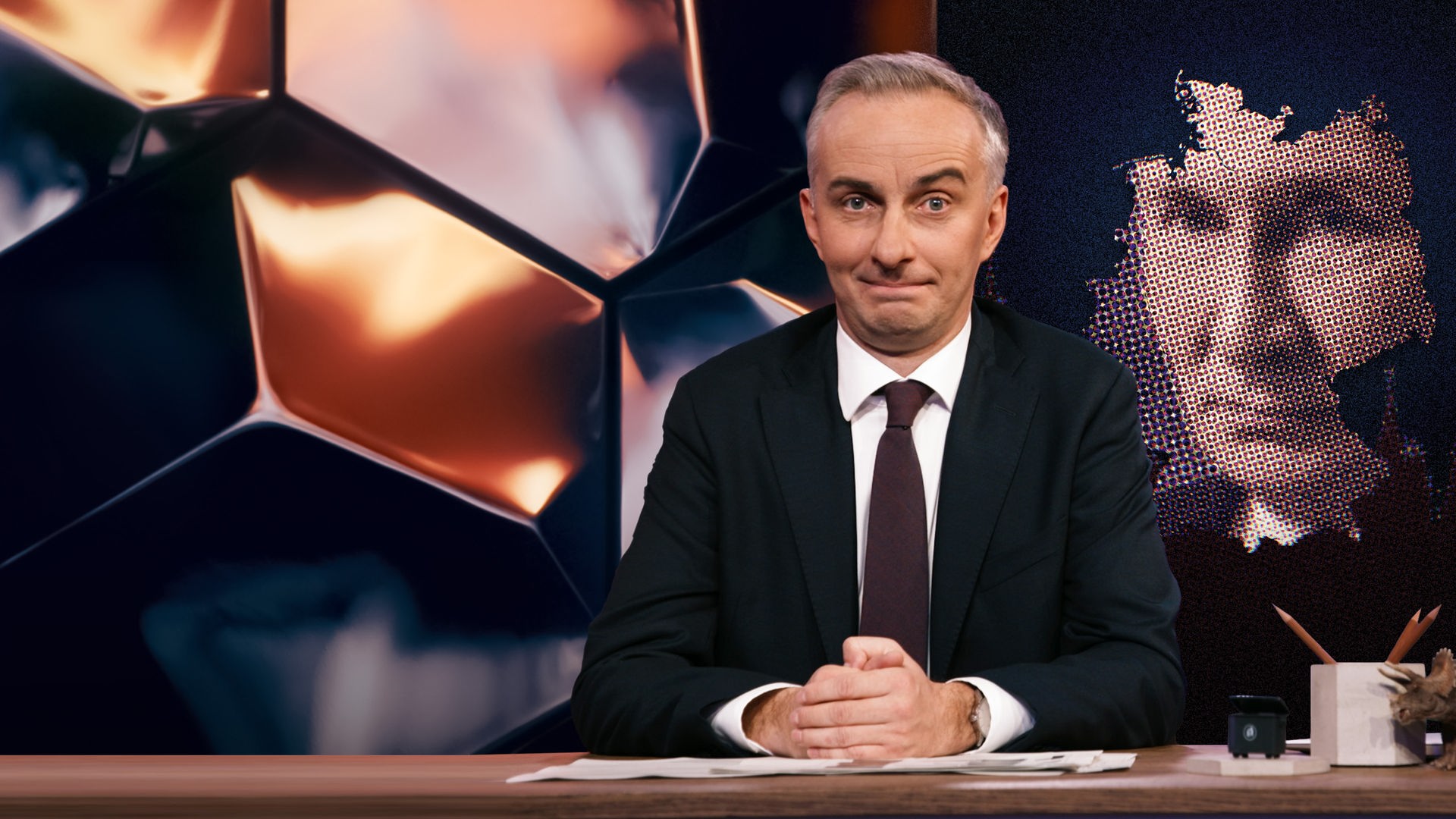 Jan Böhmermann sitzt am Schreibtisch. Rechts im Hintergrund sind die geografischen Umrisse Deutschlands abgebildet, und innerhalb dieser Umrisse befindet sich ein Bild von Putin.