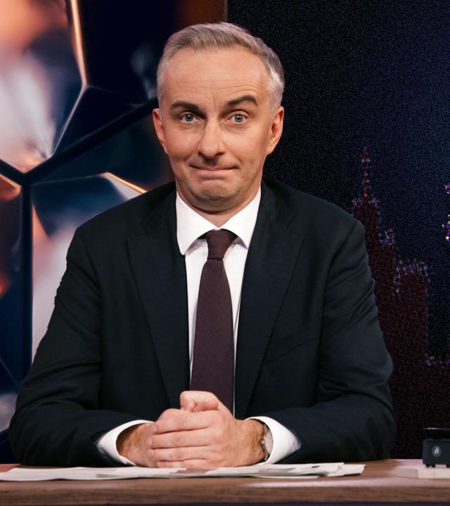 Jan Böhmermann sitzt am Schreibtisch. Rechts im Hintergrund sind die geografischen Umrisse Deutschlands abgebildet, und innerhalb dieser Umrisse befindet sich ein Bild von Putin.