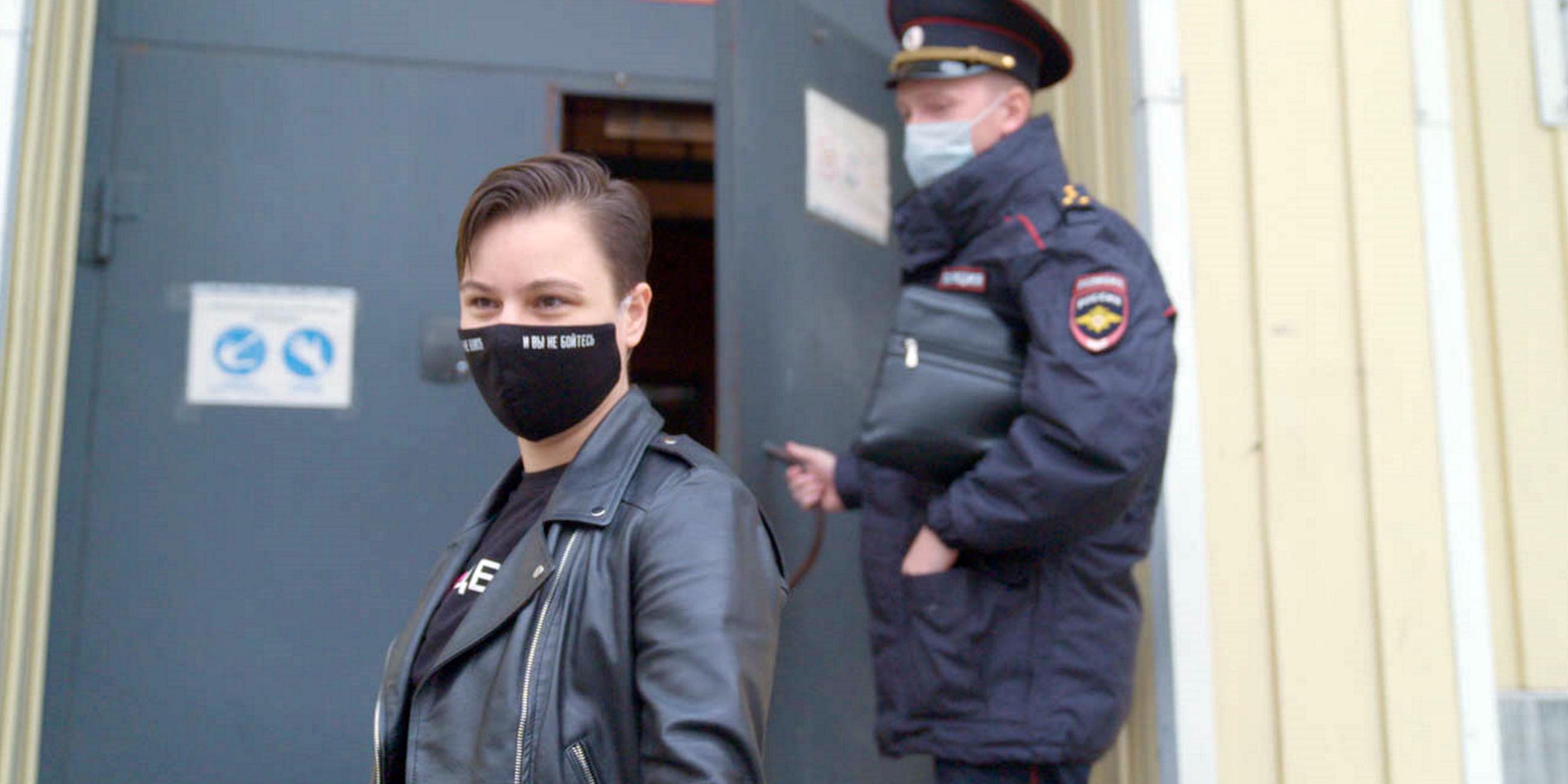 Zwei Personen stehen im Halbprofil vor der Tür zu einem Gebäude. Links eine junge Frau mit kurzem Haar, rechts ein uniformierter Polizist, der den Türgriff festhält. Beide tragen einen Mundschutz.