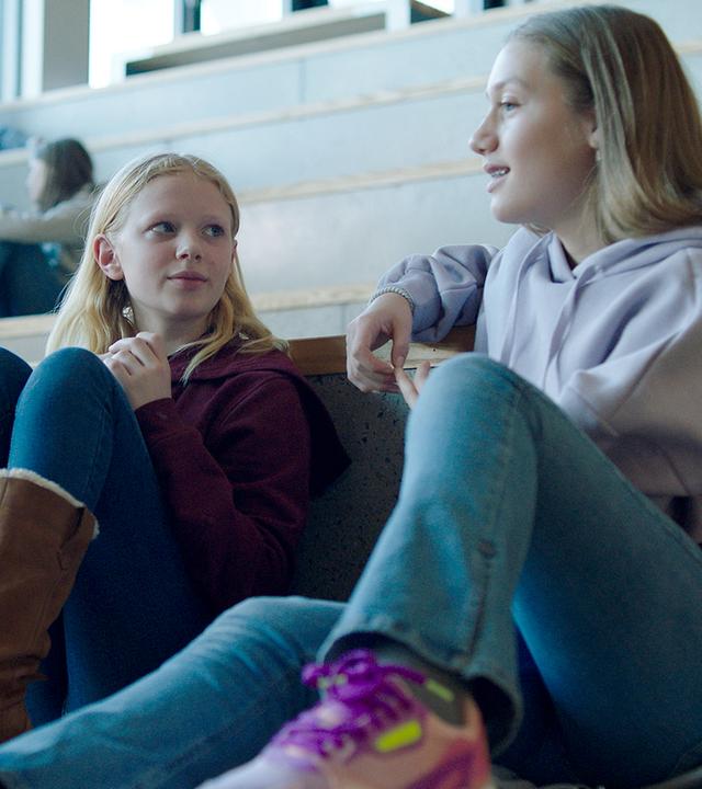 In einer Turnhalle - hell erleuchtet vom Tageslicht, das durch hochgelegene Fenster kommt - sitzen Sanne und Tara nebeneinander auf den hohen Stufen der Tribüne. Während Sanne erwartungsvoll zu Tara schaut, ist deren Blick zum Spielfeld gerichtet. Tara lächelt leicht. Beide tragen Jeans und Hoodie.