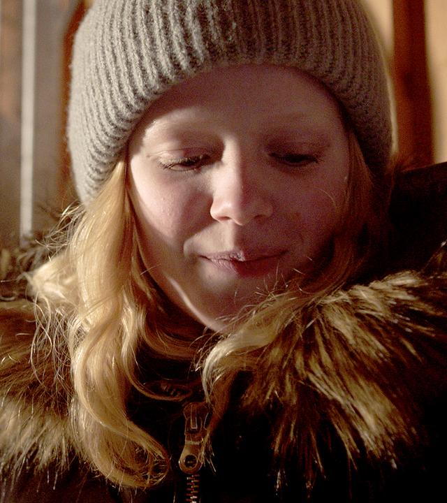 Vor einer Holzwand, an der eine Leiter hängt, stehen Daniel und Sanne mit Mützen und in dicken Jacken. Daniel schaut fragend zu Sanne, die sich schüchtern lächelnd abwendet und ihren Blick nach unten richtet.