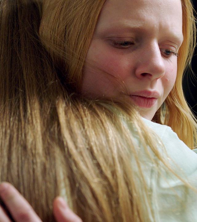 An der Eingangstür im Hausflur umarmen sich Sanne und ihre Freundin Tara. Sanne schaut besorgt über Taras Schulter. Von Tara sehen wir den Hinterkopf mit ihren langen Haaren.