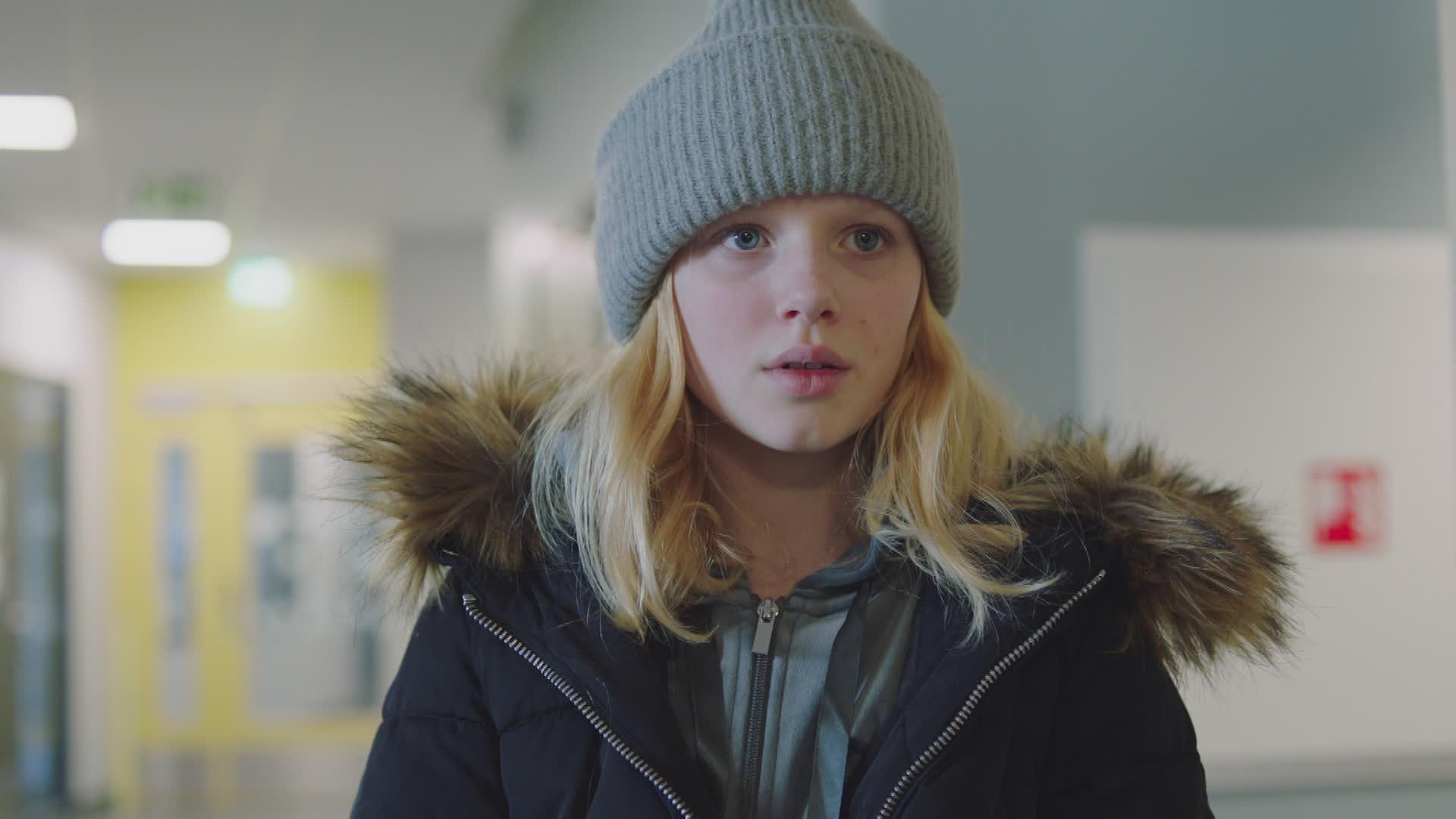 Sanne trägt eine Mütze und ihre dicke Winterjacke. Ihr Blick ist nach vorne gerichtet und sie sieht erschöpft und traurig aus.