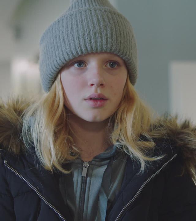 Sanne trägt eine Mütze und ihre dicke Winterjacke. Ihr Blick ist nach vorne gerichtet und sie sieht erschöpft und traurig aus.