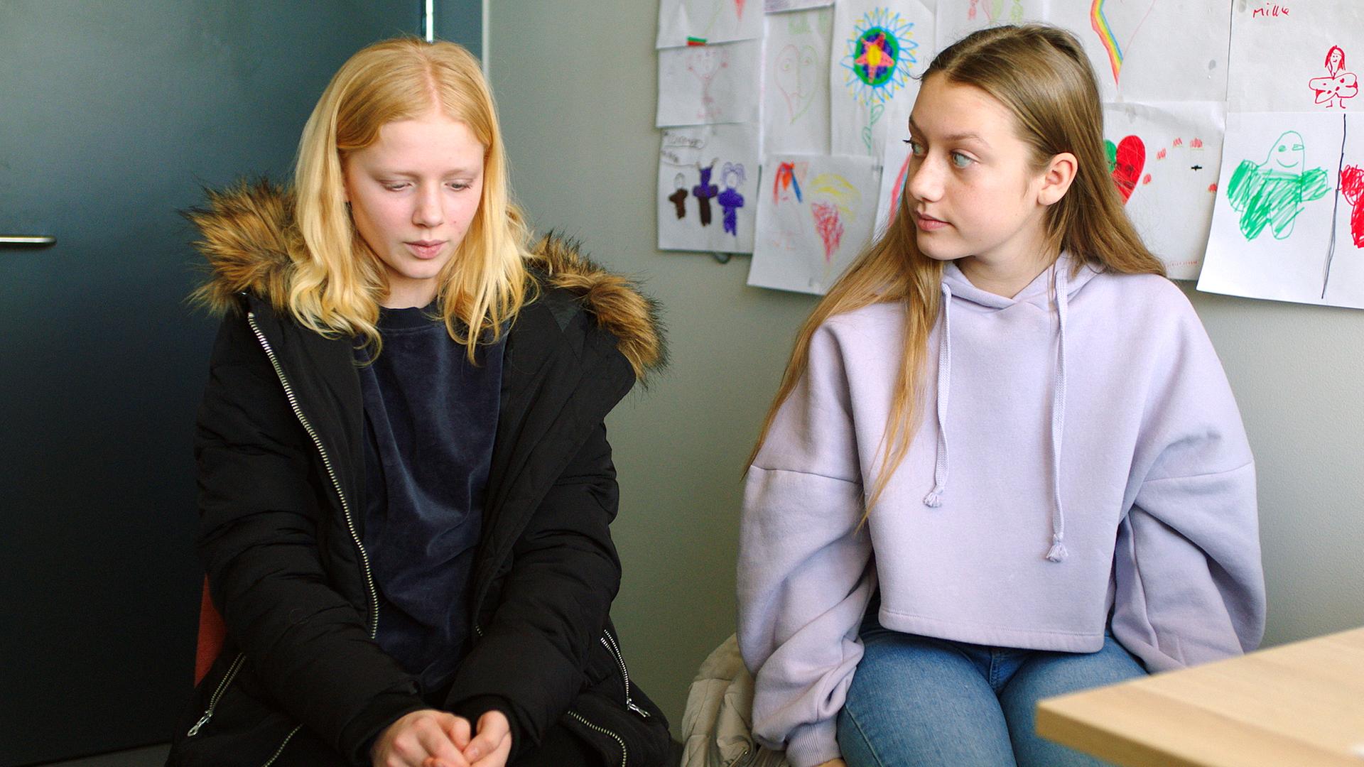 In einem Raum mit Kinderzeichnungen an einer grauen Wand sitzen Sanne und Tara nebeneinander. Sanne hat noch ihren Mantel an und schaut verlegen nach unten. Tara in einem hell-lila Hoodie schaut erwartungsvoll seitlich zu ihr. Vor ihnen ist noch eine Ecke eines Tisches zu erkennen.