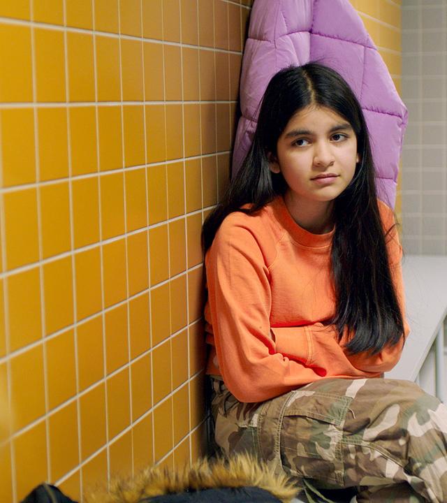 In einer gelb-gekachelten Umkleidekabine sitzen Michelle und Sanne weit auseinander auf einer grauen Bank, die an der Wand steht. Michelle schaut mit ernster Miene und vor dem Bauch verschränkten Armen zu Sanne, die von hinten zu sehen ist.