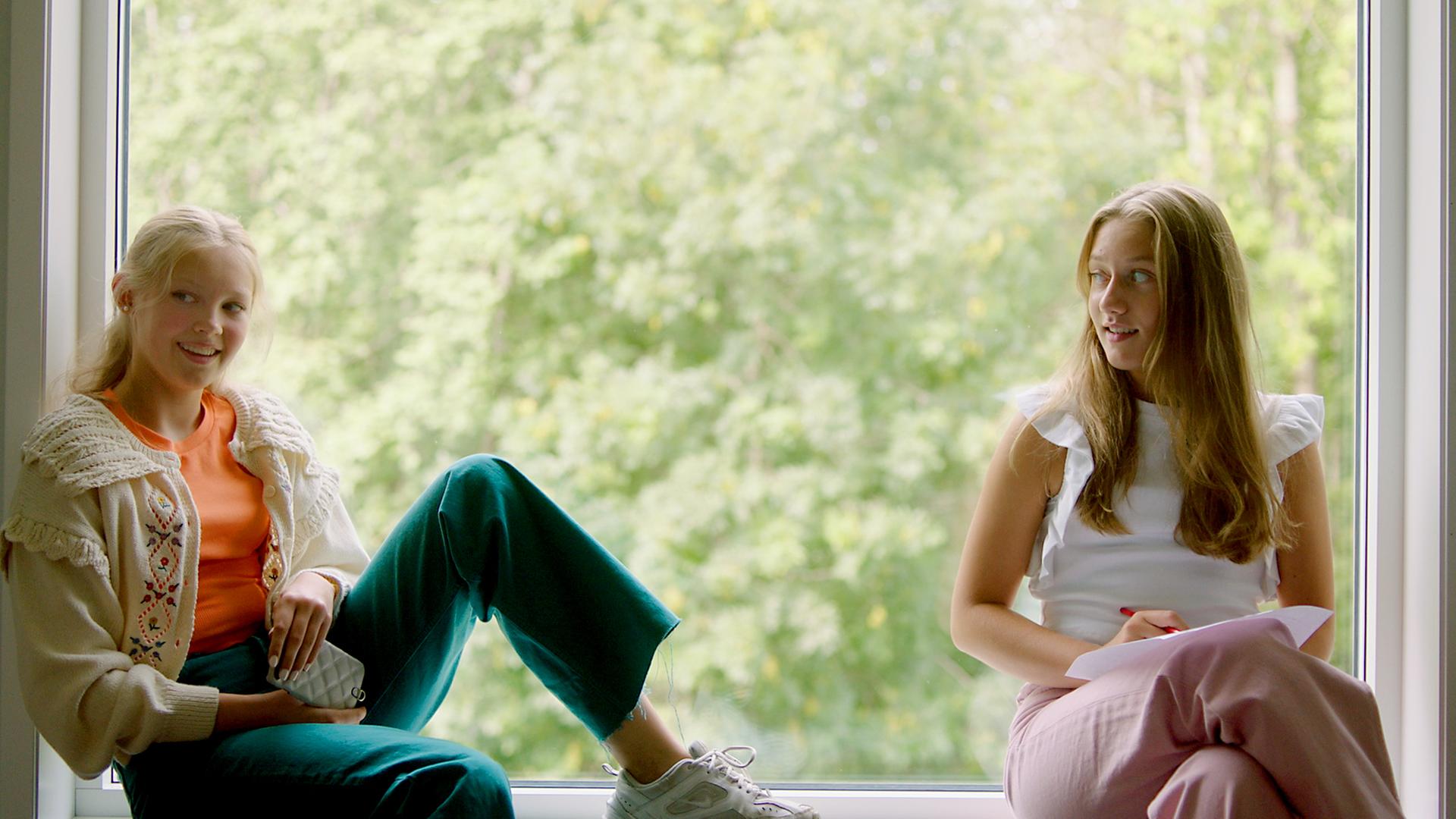 Tara und Mina sitzen vor einem großen Fenster auf einer Fensterbank. Mina sitzt quer und hat einen Fuß mit angewinkeltem Bein auf dem Fensterbrett abgestellt. Dabei schaut sie offen lächelnd nach vorne in den Raum. Tara hingegen sitzt mit überschlagenem Bein eng am Rahmen und schaut verhalten-lächelnd seitlich zu Mina. Mina hält ein Handy in ihren Händen und Tara einen Zettel und einen Stift. Draußen, durch das Fenster, sind hellgrüne Bäume im Tageslicht zu sehen.
