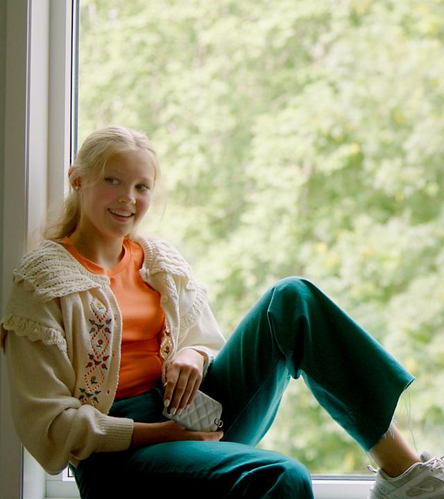 Tara und Mina sitzen vor einem großen Fenster auf einer Fensterbank. Mina sitzt quer und hat einen Fuß mit angewinkeltem Bein auf dem Fensterbrett abgestellt. Dabei schaut sie offen lächelnd nach vorne in den Raum. Tara hingegen sitzt mit überschlagenem Bein eng am Rahmen und schaut verhalten-lächelnd seitlich zu Mina. Mina hält ein Handy in ihren Händen und Tara einen Zettel und einen Stift. Draußen, durch das Fenster, sind hellgrüne Bäume im Tageslicht zu sehen.