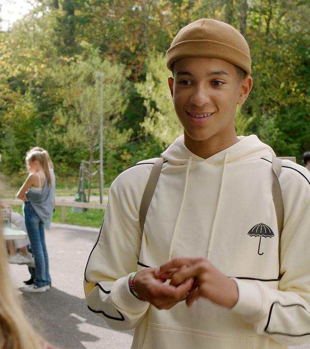 Bei Sonnenlicht auf dem Schulhof stehen sich Jonas und Tara gegenüber. Jonas schaut Tara freundlich lächelnd an. Tara ist nur von hinten zu sehen. Jonas hält seine Hände vor den Oberkörper und spielt mit seinen Fingern. Im Hintergrund sind andere Kinder zu sehen.