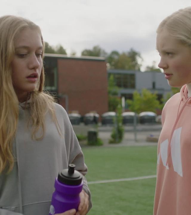 Bei Tageslicht stehen Tara und Mina sich gegenüber auf einem Wiesen-Sportplatz - im Hintergrund ist das Schulgebäude. Mina guckt zweifelnd in Taras Gesicht, während Tara ernst nach unten schaut, also ob sie Minas Blick ausweicht.