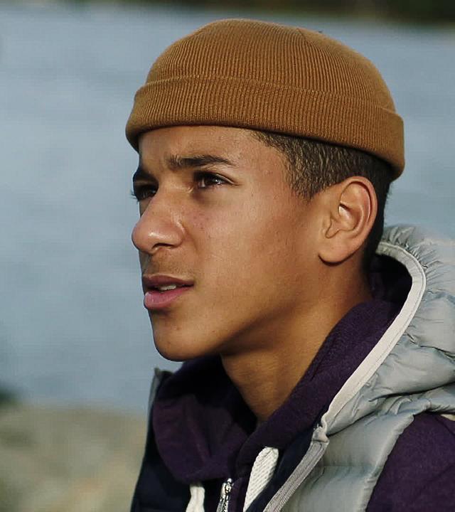 Bei Tageslicht sitzen Jonas und Tara nebeneinander am Wasser. Jonas trägt eine dicke Jacke zusätzlich mit Daunenweste darüber und eine Mütze auf dem Kopf. Er schaut mit ernstem Gesicht seitlich nach oben, während Tara ihn anschaut.