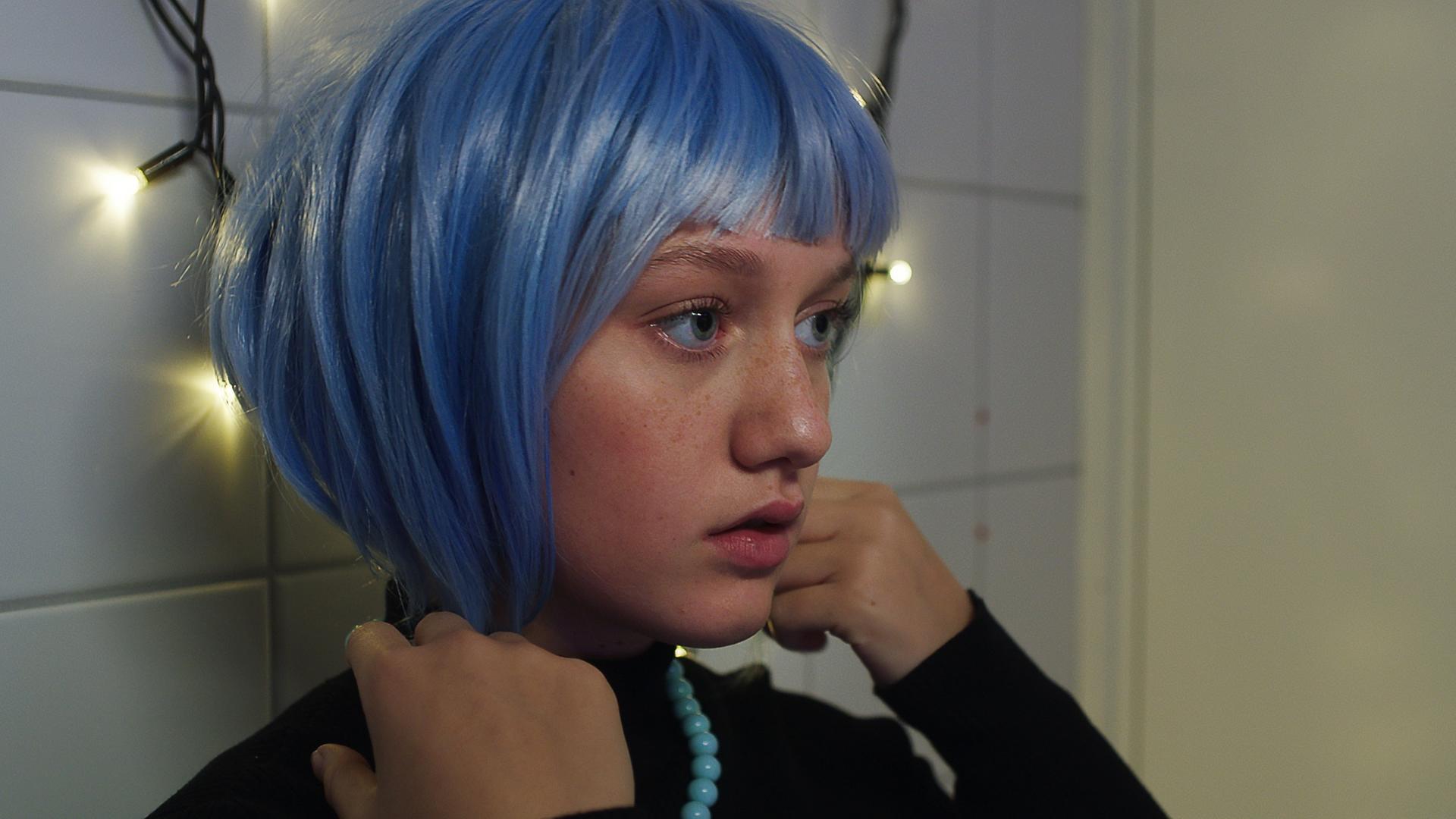 Vor einer weißen Kachelwand, die mit einer leuchtenden Lichterkette dekoriert ist, steht Tara. Sie trägt eine blaue Perücke mit Bob-Frisur auf dem Kopf. Ihre Hände hält sie rechts und links an die unteren Haarsträhnen. Sie schaut ernst und etwas fragend nach vorne - schätzungsweise in einen Spiegel.