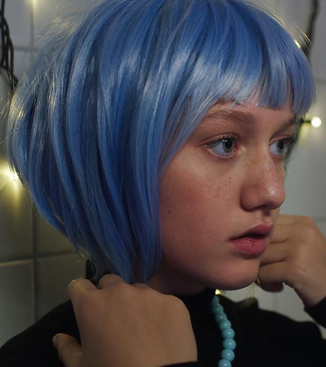 Vor einer weißen Kachelwand, die mit einer leuchtenden Lichterkette dekoriert ist, steht Tara. Sie trägt eine blaue Perücke mit Bob-Frisur auf dem Kopf. Ihre Hände hält sie rechts und links an die unteren Haarsträhnen. Sie schaut ernst und etwas fragend nach vorne - schätzungsweise in einen Spiegel.