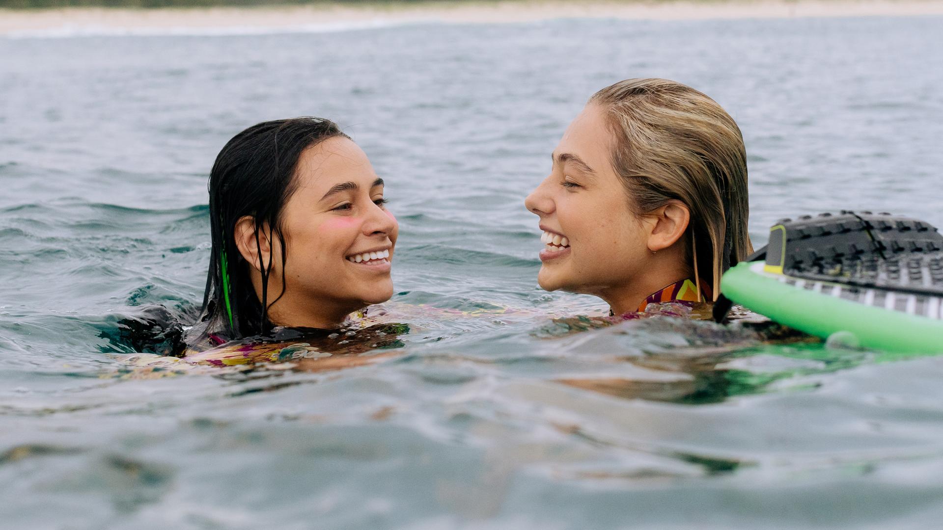 Summer und Wren befinden sich gemeinsam im Wasser und nur ihre Köpfe sind zu sehen .Sie lachen fröhlich. Neben ihnen befindet sich ein grünes Surfbrett.