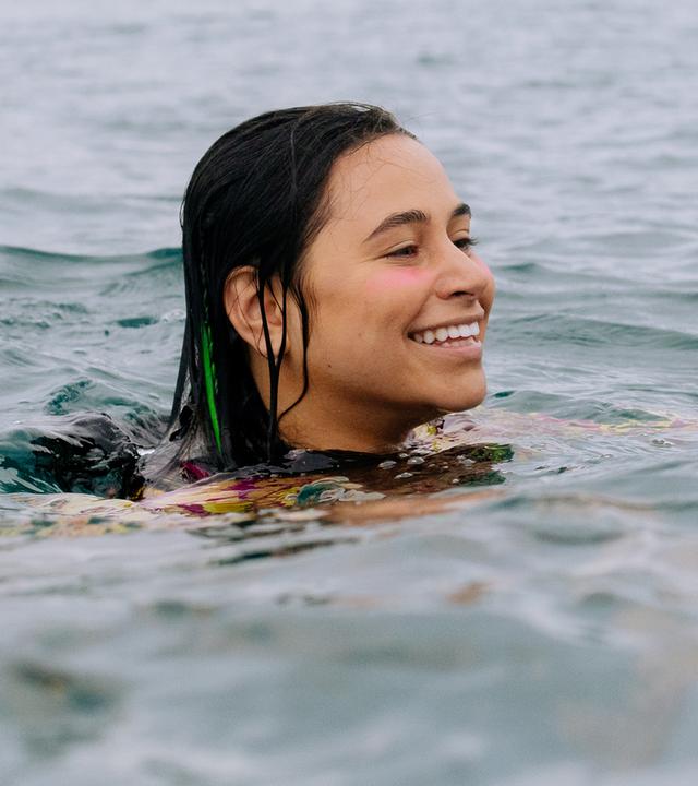 Summer und Wren befinden sich gemeinsam im Wasser und nur ihre Köpfe sind zu sehen .Sie lachen fröhlich. Neben ihnen befindet sich ein grünes Surfbrett.