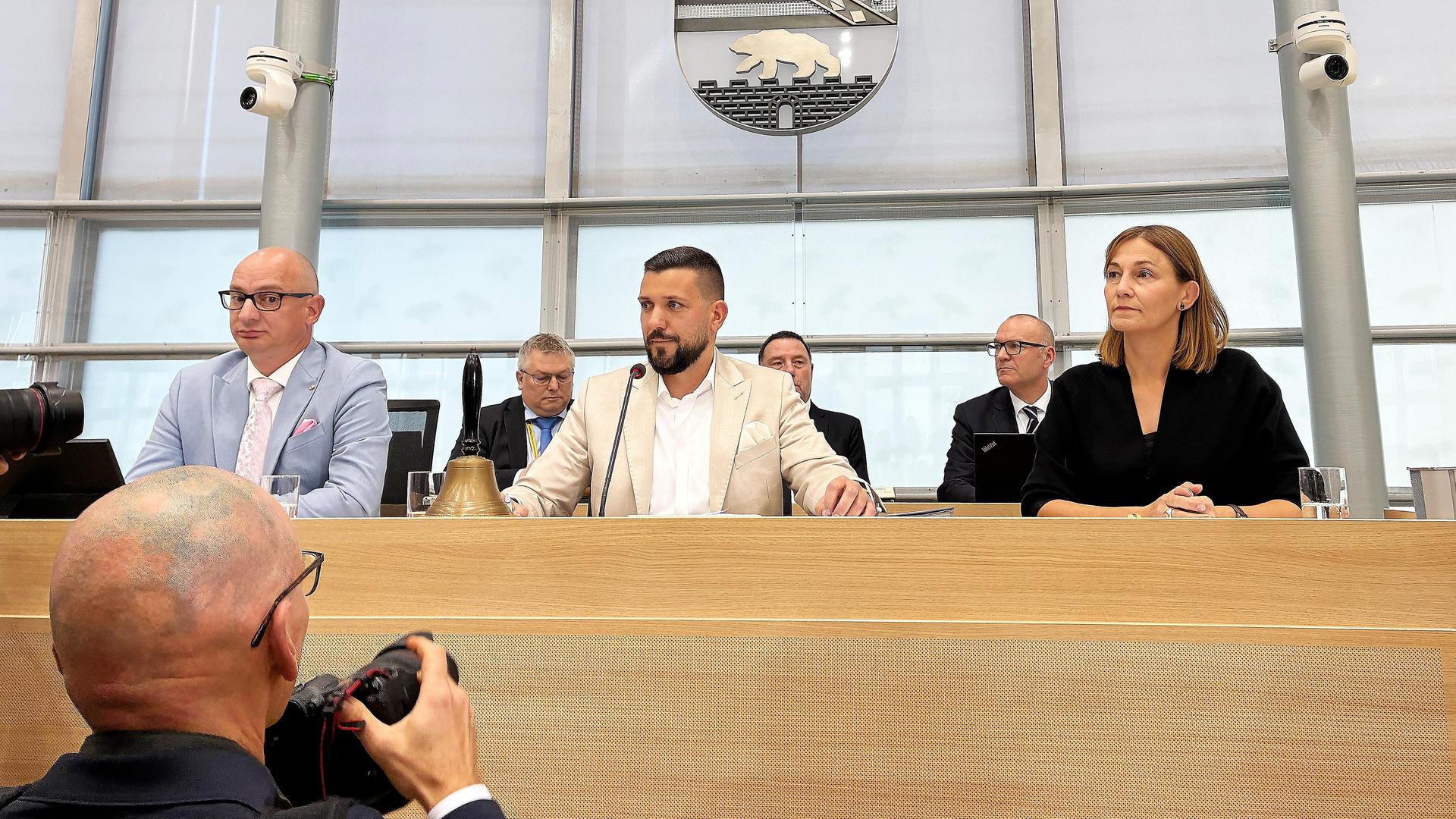 Hinter einem holzverkleideten Podest sitzen zwei Männer und eine Frau, hinter ihnen ist eine weitere Reihe mit Männern in Anzügen zu sehen. Über allen hängt das Wappen von Sachsen-Anhalt.