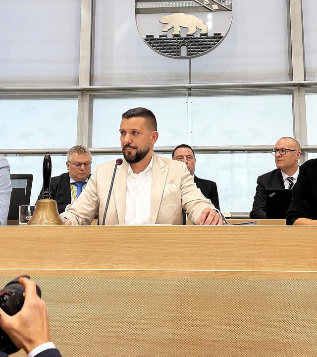 Hinter einem holzverkleideten Podest sitzen zwei Männer und eine Frau, hinter ihnen ist eine weitere Reihe mit Männern in Anzügen zu sehen. Über allen hängt das Wappen von Sachsen-Anhalt.